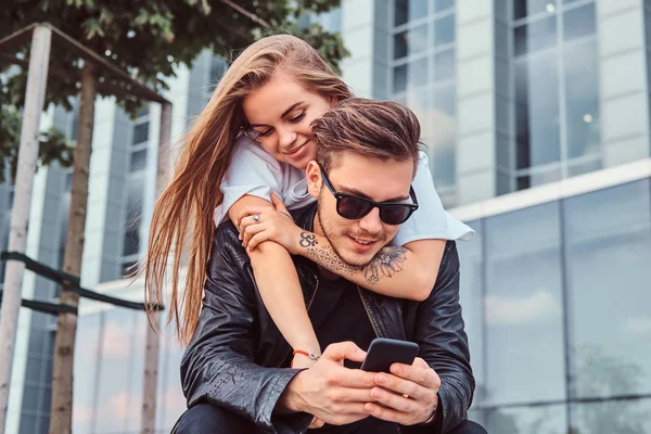 Attraktive trendy gekleidete junge Paar sitzt zusammen auf Bank in der Nähe von Wolkenkratzer - hübsches Mädchen umarmt ihren Freund. — Stockfoto