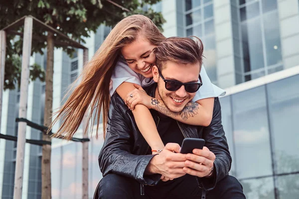 Aantrekkelijke trendy gekleed jonge paar samen zitten op bankje in de buurt van wolkenkrabber - mooi meisje omarmt haar vriendje. — Stockfoto