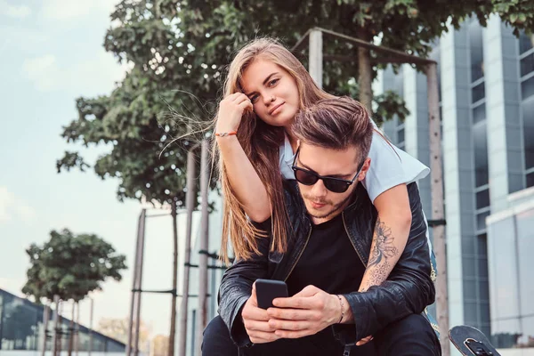 Aantrekkelijke trendy gekleed jonge paar samen zitten op bankje in de buurt van wolkenkrabber - mooi meisje omarmt haar vriendje. — Stockfoto