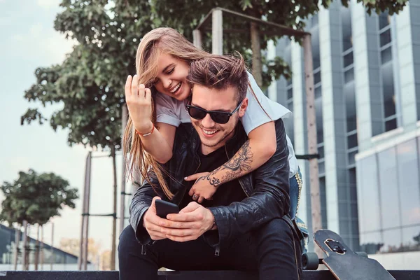 Attractive trendy dressed young couple sitting together on bench near skyscraper - pretty girl embraces her boyfriend. — Stock Photo, Image