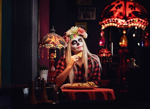 Young blonde girl with undead makeup in flower wreath eating nachos at a mexican restaurant. — Stock Photo, Image