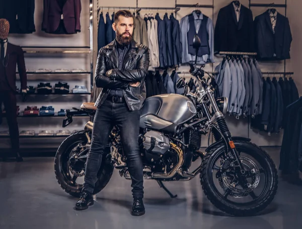 Brutaler Mann in schwarzer Jacke lehnt an einem Retro-Sportmotorrad im Herrenbekleidungsgeschäft. — Stockfoto