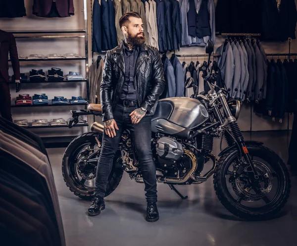 Brutal male dressed in black jacket leaning on a retro sports motorbike at the mens clothing store. — Stock Photo, Image