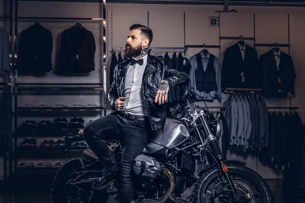 Стильный татуированный бородатый мужчина в черной кожаной куртке и галстуке-бабочке позирует рядом с ретро спортивным мотоциклом в магазине мужской одежды . — стоковое фото