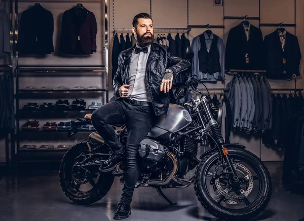 Stylish tattooed bearded man with dressed in black leather jacket and bow tie posing near retro sports motorbike at mens clothing store. — Stock Photo, Image