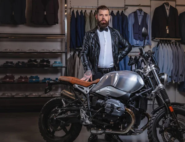 Homem barbudo tatuado elegante com vestido de jaqueta de couro preto e gravata borboleta posando perto de moto retro esportes na loja de roupas dos homens . — Fotografia de Stock