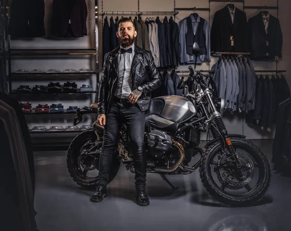 时尚纹身胡子男子身着黑色皮革夹克和弓领带摆在男装服装店附近复古运动摩托车. — 图库照片