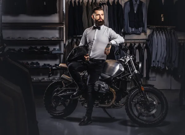 Elegantly dressed tattooed male with stylish beard holds black leather jacket posing near retro sports motorbike at the mens clothing store. — Stock Photo, Image