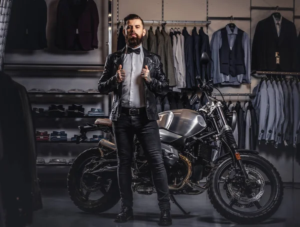 Stylish tattooed bearded man with dressed in black leather jacket and bow tie posing near retro sports motorbike at mens clothing store. — Stock Photo, Image