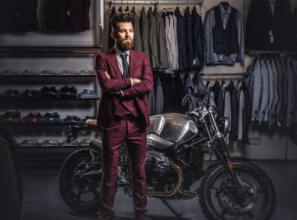 英俊的男子与时尚的胡须和头发身着复古红色西装摆在男装服装店附近复古运动摩托车. — 图库照片
