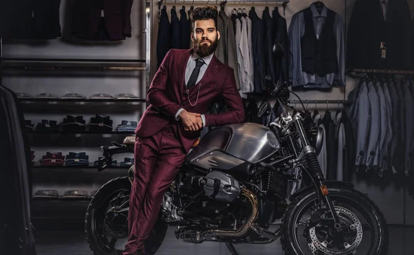 Handsome man with a stylish beard and hair dressed in vintage red suit posing near retro sports motorbike at mens clothing store. — Stock Photo, Image