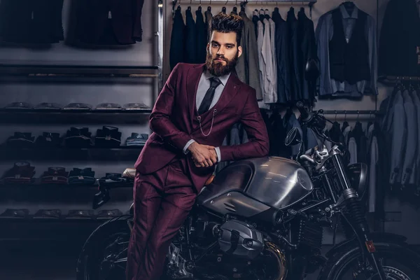 Handsome man with a stylish beard and hair dressed in vintage red suit posing near retro sports motorbike at mens clothing store. — Stock Photo, Image