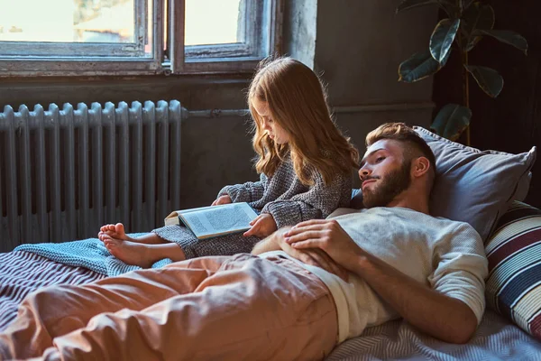 Bonito jovem pai e filha lendo o livro de histórias juntos enquanto estavam deitados na cama . — Fotografia de Stock