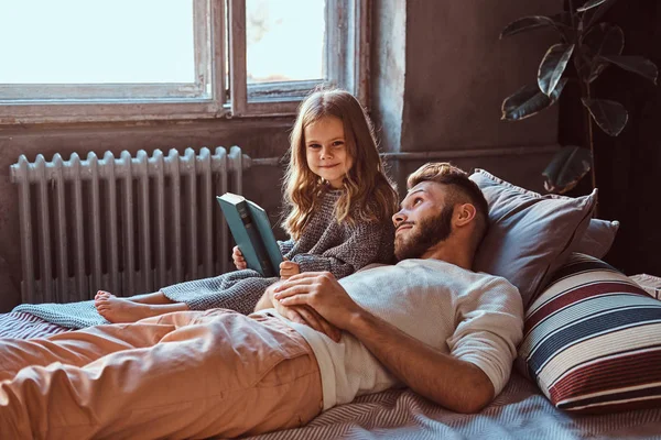 Guapo joven padre e hija leyendo cuentos juntos mientras yacen en la cama . — Foto de Stock