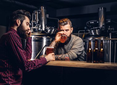 Two stylish bearded hipsters friends drinking craft beer together at the indie brewery. clipart