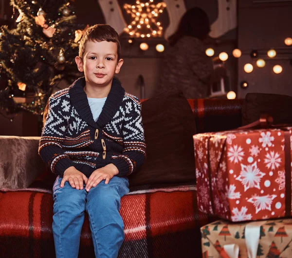 Niedlicher Junge im Pullover sitzt auf Sofa in der Nähe von Geschenkboxen in dekoriertem Raum zur Weihnachtszeit. — Stockfoto