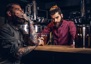 Tattooed male with stylish beard and hair holds pint of craft beer sitting at the bar counter in the indie brewery. clipart