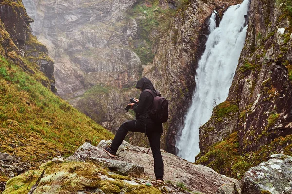 Fotografo naturalistico turista con macchina fotografica scatta mentre in piedi sulla montagna contro una cascata . — Foto Stock