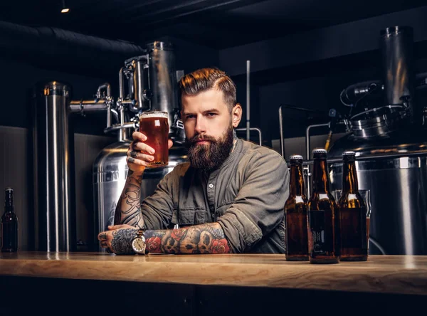 Hombre hipster tatuado con barba elegante y cabello bebiendo cerveza sentado en el mostrador del bar en la fábrica de cerveza indie . — Foto de Stock