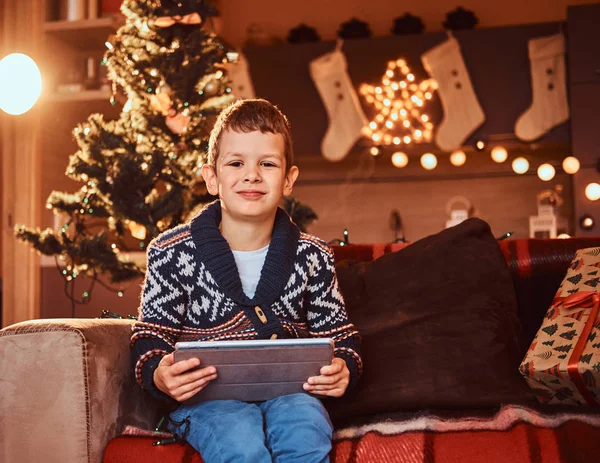 Счастливый симпатичный мальчик в свитере держит подарочную тарелку и смотрит в камеру, сидя на диване в украшенной комнате на Рождество . — стоковое фото