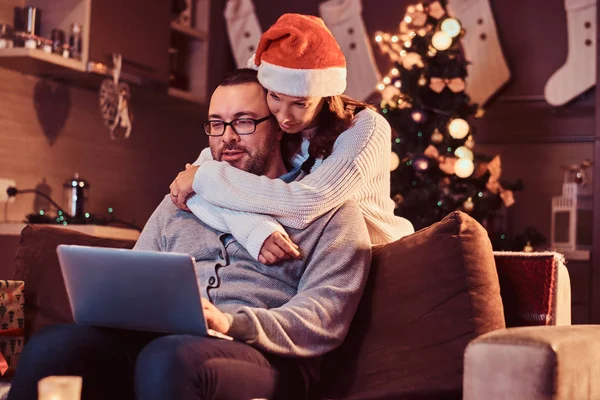 一对可爱的情侣迷人的妇女在圣诞老人的帽子拥抱她的男人和使用笔记本电脑的肖像. — 图库照片