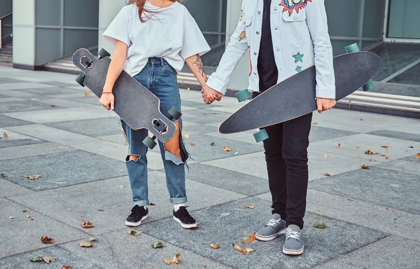 Foto recortada de jóvenes patinadores vestidos de moda tomados de la mano mientras están de pie cerca de rascacielos . —  Fotos de Stock
