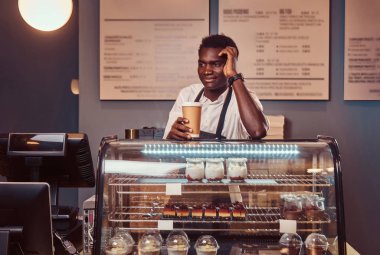 Bir fincan kahve ve kahve dükkanında iş günü sonra rahatlatıcı bir yakışıklı Afrika barista beyaz gömlek ve önlük portresi tutar.