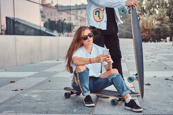 Jonge hipster meisje zittend op een longboard en met behulp van een smartphone terwijl haar vriendje permanent in de buurt van. — Stockfoto