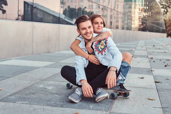 Trendy gekleidetes junges Paar - hübsches Mädchen umarmt ihren Freund, während sie zusammen auf einem Skateboard in der Nähe eines Wolkenkratzers sitzen. — Stockfoto