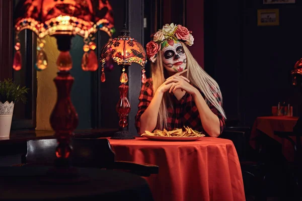 Young blonde girl with undead makeup in flower wreath eating nachos at a mexican restaurant. — Stock Photo, Image