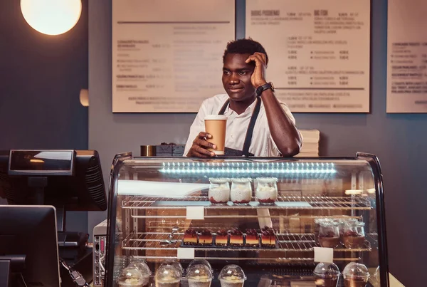 Retrato de un guapo barista africano en camisa blanca y delantal sostiene una taza de café y relajarse después del día de trabajo en la cafetería . —  Fotos de Stock