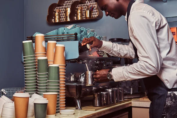 Πλευρά προβολή της αφρικανικής αμερικανικής barista στο λευκό πουκάμισο και ποδιά κάνει καφέ με μια μηχανή του καφέ. — Φωτογραφία Αρχείου