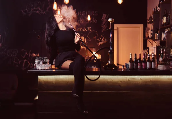 Menina morena sexy em roupas pretas sedutoras fuma um narguilé enquanto está sentado no balcão em uma boate ou bar . — Fotografia de Stock