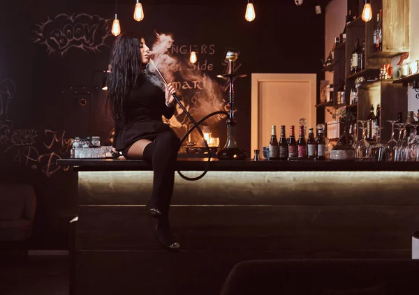 Sexy fille brune dans des vêtements noirs séduisants fume un narguilé tout en étant assis sur le comptoir dans une boîte de nuit ou un bar . — Photo