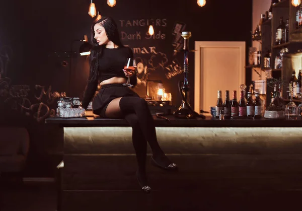매혹적인 검은 옷에 뜨거운 갈색 머리 여자는 나이트 클럽에 카운터에 앉아있는 동안 와인 글라스를 보유 하. — 스톡 사진