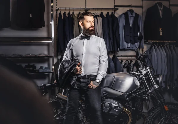 Elegantly dressed tattooed male with stylish beard holds black leather jacket posing near retro sports motorbike at the mens clothing store. — Stock Photo, Image