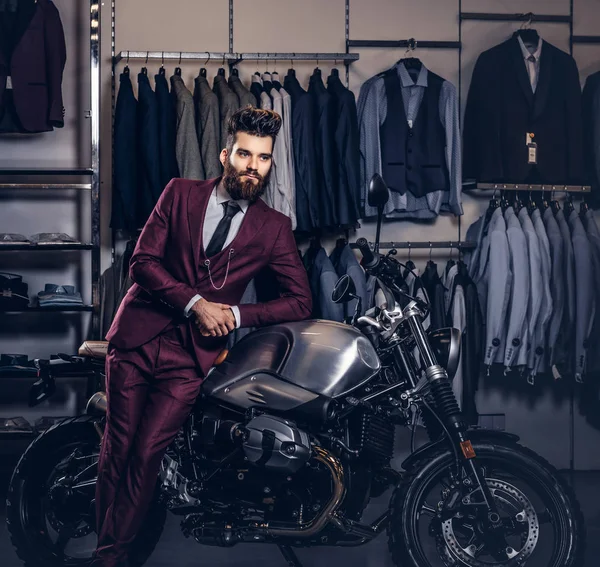 英俊的男子与时尚的胡须和头发身着复古红色西装摆在男装服装店附近复古运动摩托车. — 图库照片