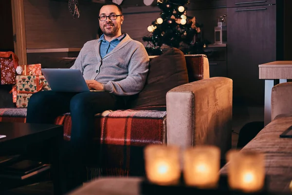 Hombre feliz en gafas vestidas con suéter cálido sostener el ordenador portátil y mirando a la cámara mientras está sentado en el sofá en tiempo de Navidad . — Foto de Stock