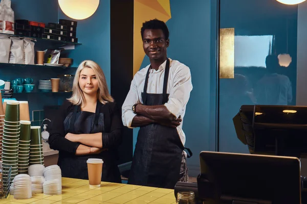 Dos jóvenes baristas multirraciales en delantales de pie en su cafetería de moda . —  Fotos de Stock