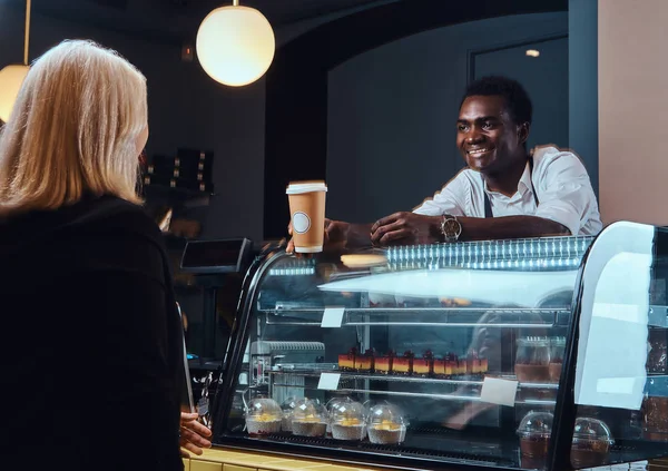 Smiling African American barista in uniform giving a cup of coffee to his client at the trendy coffee shop.