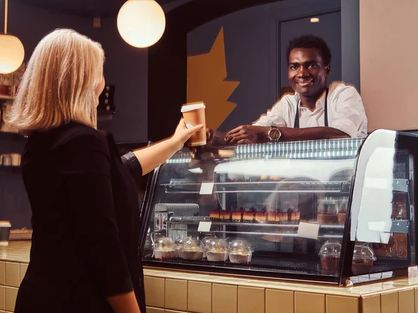 Barista afroamericano sonriente en uniforme dando una taza de café a su cliente en la cafetería de moda . —  Fotos de Stock