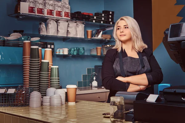 Beautiful experienced female barista in apron standing with crossed arms at the coffee shop.