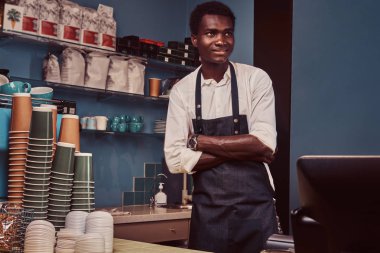 Trendy bir kahve dükkanı kontuarında çapraz kollu yakışıklı Afrika barista ayakta portresi.
