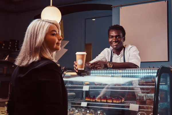 Barista afroamericano sonriente en uniforme dando una taza de café a su cliente en la cafetería de moda . —  Fotos de Stock
