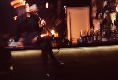 Baştan çıkarıcı siyah giysili sıcak esmer kızı bir gece kulübünde sayaçta otururken bir nargile içiyor.