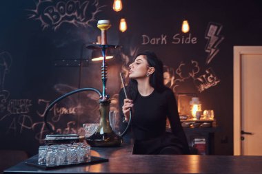 Baştan çıkarıcı siyah giysili sıcak esmer kızı bir gece kulübünde tezgahın üstünde otururken bir nargile içiyor.