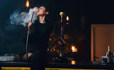 Seksi esmer kız baştan çıkarıcı siyah giysili bir gece kulübü ya da bar counter üzerinde otururken bir nargile içiyor.