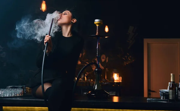 Szexi barna lány, csábító fekete ruhában dohányzik a vízipipa ülve, a számláló egy szórakozóhely vagy a bárban. — Stock Fotó