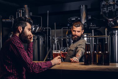 Two stylish bearded hipsters friends drinking craft beer together at the indie brewery. clipart