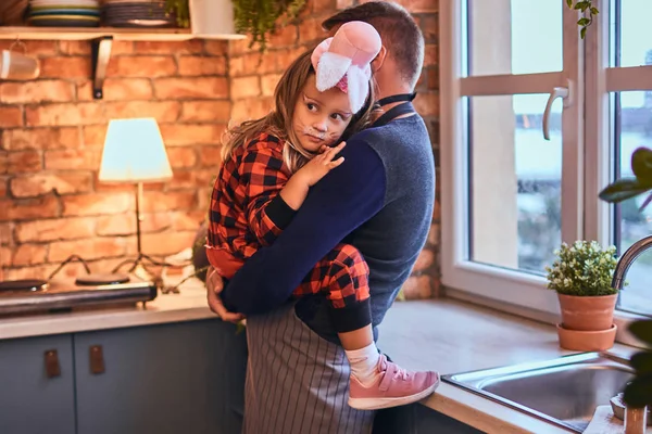 Niedliches kleines Mädchen in Kaninchen-Make-up und Hut sitzt auf den Händen seines Vaters in der Loft-Küche am Morgen. — Stockfoto